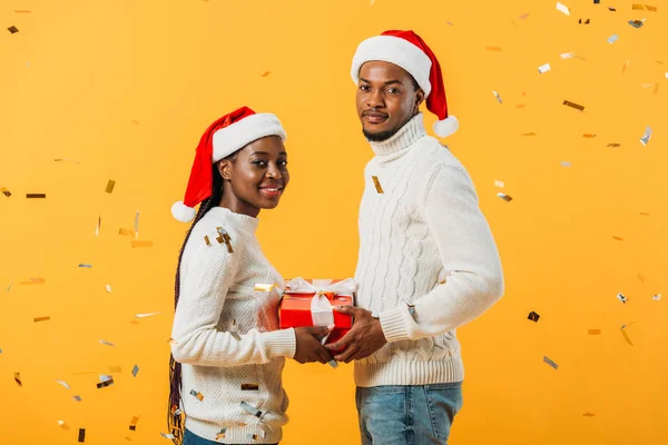 side view of African American couple in Santa hats holding gift box on yellow background with confetti