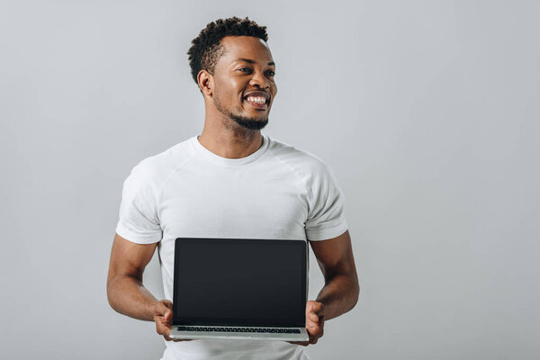 Афроамериканец улыбается и показывает ноутбук с чистым экраном и смотрит в сторону изолированных на сером

