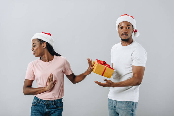 African American woman in Santa hat rejecting gift box from man isolated on grey