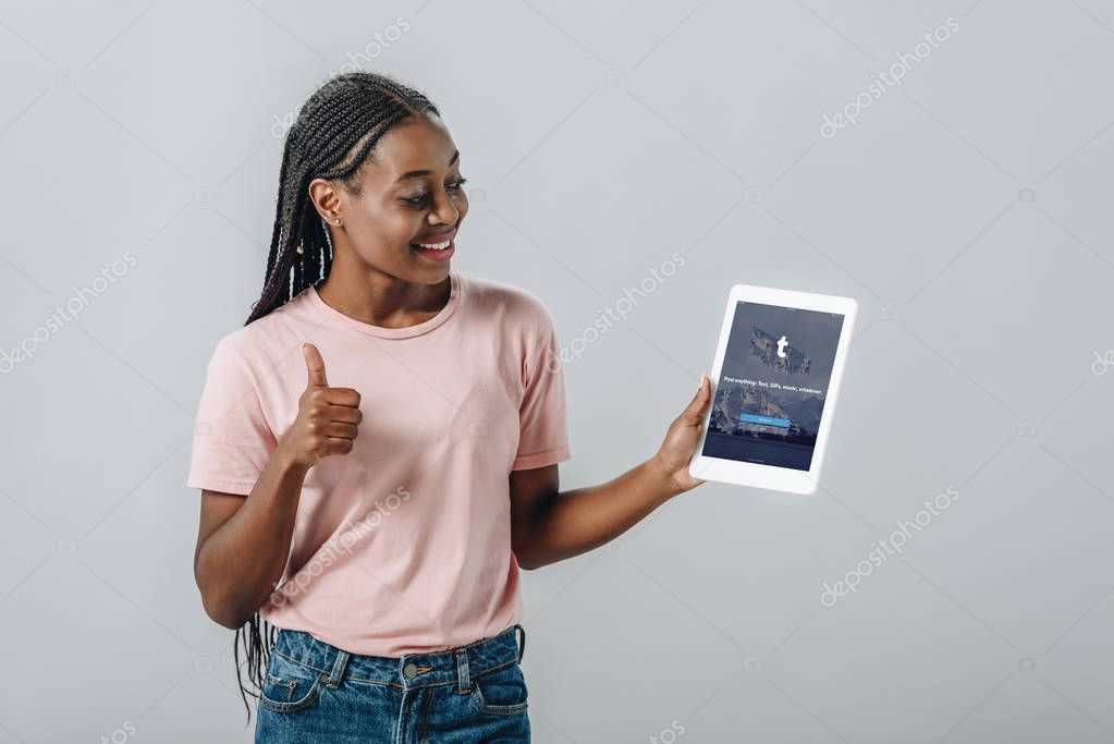 KYIV, UKRAINE - AUGUST 2, 2019: African American woman holding digital tablet with tumblr app and showing thumb up isolated on grey