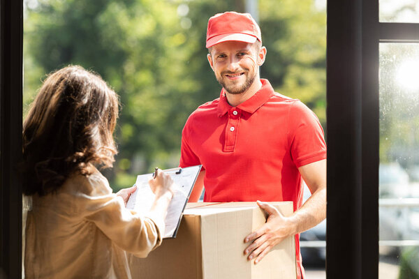 happy delivery man holding box near woman with clipboard and pen 