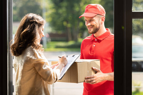 happy delivery man holding box and looking at woman with clipboard and pen 