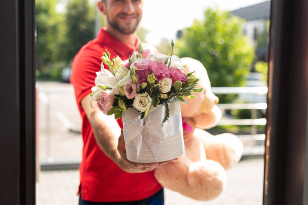cropped view of bearded delivery man holding teddy bear and flowers 