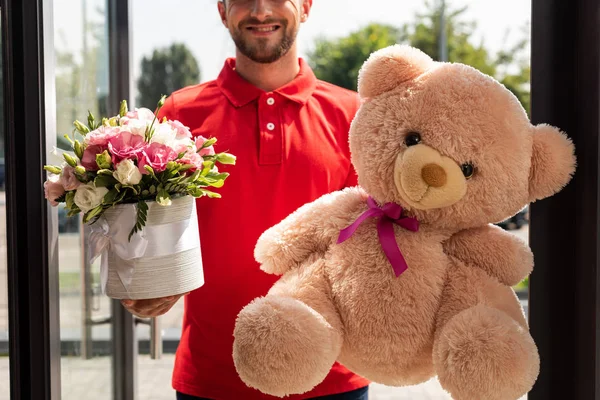 cropped view of positive delivery man holding teddy bear and flowers