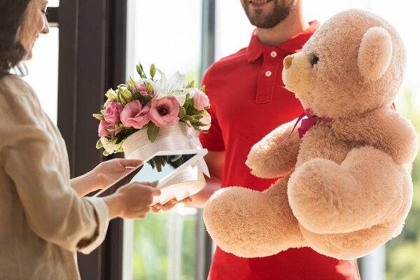 cropped view of happy delivery man holding teddy bear and flowers near girl with digital tablet 