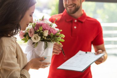 selective focus of woman smelling flowers near delivery man holding clipboard clipart