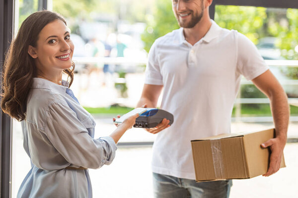 selective focus of happy woman paying while holding smartphone near credit card reader in hand on delivery man 