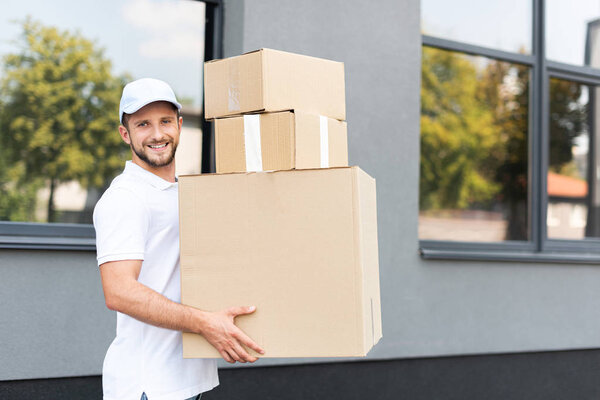 cheerful delivery man in cap looking at camera and holding boxes 