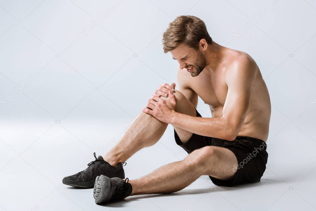 bearded shirtless sportsman with knee pain on grey