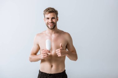 front view of smiling shirtless sportsman holding ointment and showing thumb up isolated on grey clipart