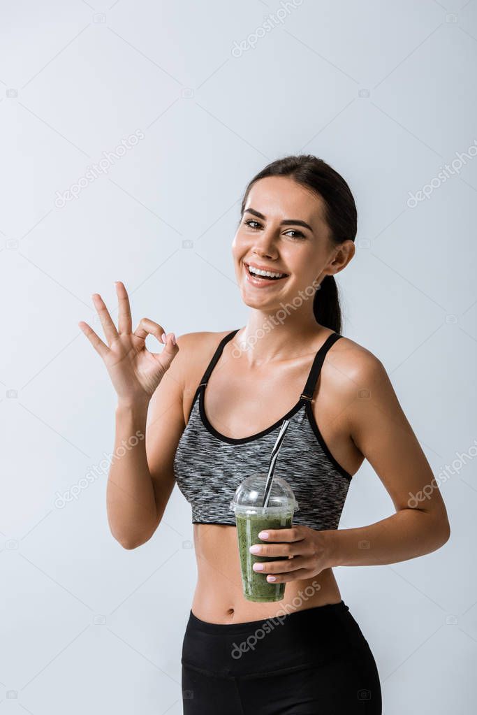 smiling sportswoman holding smoothie and showing okay sign isolated on grey