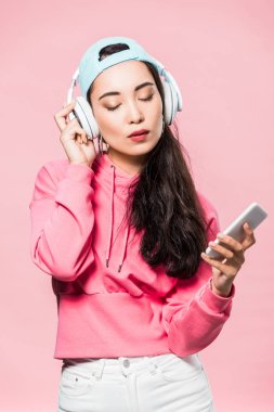 Çekici Asyalı kadın kazak ve şapka takıyor müzik dinliyor ve pembe üzerine izole edilmiş akıllı telefon tutuyor. 