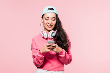 Kazaklı ve şapkalı gülümseyen Asyalı kadın pembe üzerine izole edilmiş akıllı telefon tutan kulaklıklı. 
