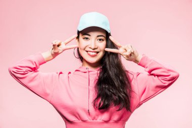 Pembe kazaklı, şapkalı, gülümseyen ve pembe üzerine izole edilmiş barış işareti gösteren çekici Asyalı kadın. 