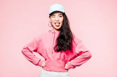 attractive asian woman in pink pullover and cap winking and sticking out tongue isolated on pink  clipart