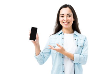 Çekici ve gülümseyen, kot gömlekli Asyalı kadın. Beyazlar içinde izole edilmiş akıllı telefonu işaret ediyor. 