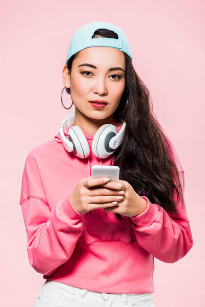 attractive asian woman in pullover and cap with headphones holding smartphone isolated on pink 
