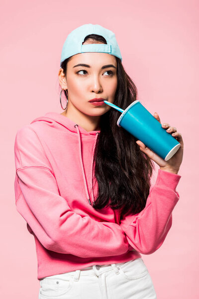 задумчивая азиатская женщина в пуловере и колпачок пить из пластиковой чашки изолированы на розовый
 