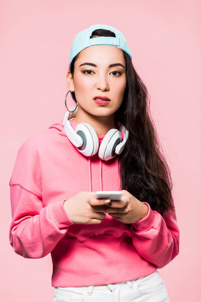 skeptical asian woman in pullover and cap with headphones holding smartphone isolated on pink 