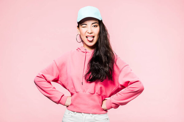 attractive asian woman in pink pullover and cap winking and sticking out tongue isolated on pink 