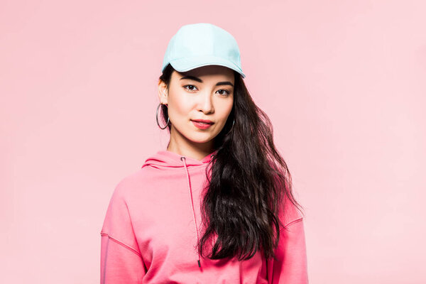 attractive asian woman in pink pullover and cap looking at camera isolated on pink 