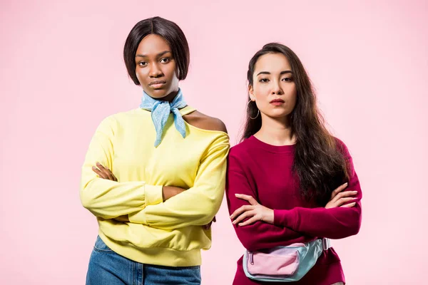 attractive asian and african american friends with crossed arms looking at camera isolated on pink