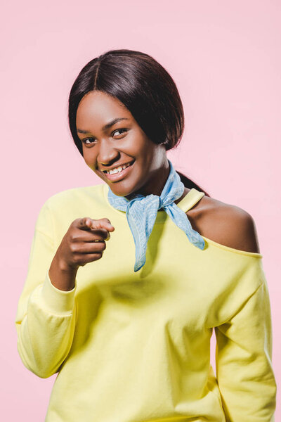 smiling african american woman in yellow pullover and scarf pointing with finger isolated on pink 
