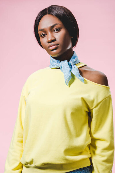african american woman in yellow pullover and scarf looking at camera isolated on pink 