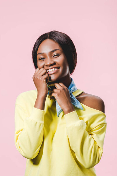 smiling african american woman in yellow pullover and scarf looking at camera isolated on pink 