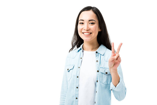 привлекательная и улыбающаяся азиатка в джинсовой рубашке, показывающая знак мира, изолированный на белом
 