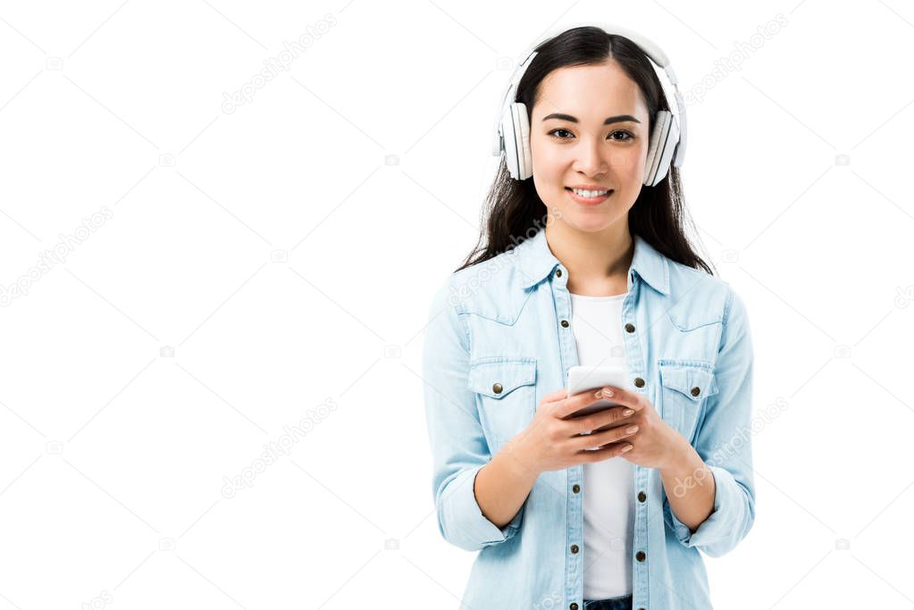 attractive and smiling asian woman in denim shirt listening music and holding smartphone isolated on white 