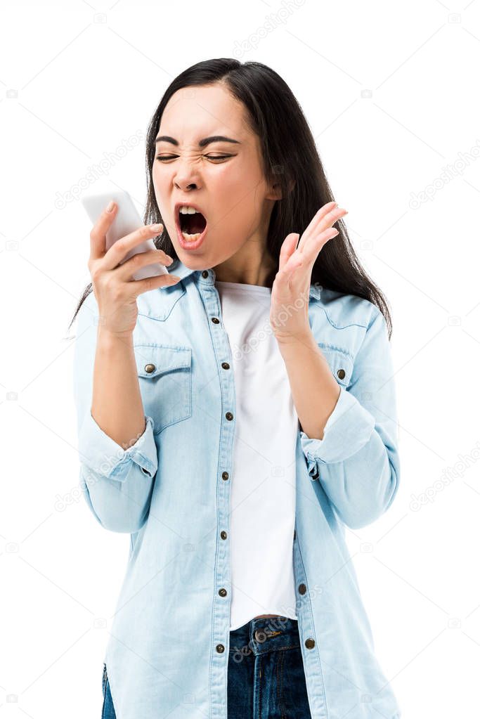 attractive and angry asian woman in denim shirt screaming and holding smartphone isolated on white 