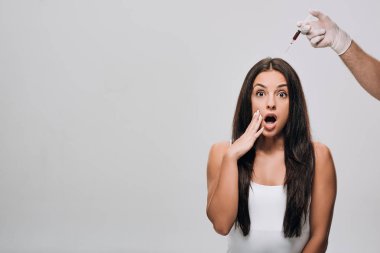 cosmetologist in latex gloves doing scalp mesotherapy to shocked beautiful woman with long hair isolated on grey clipart