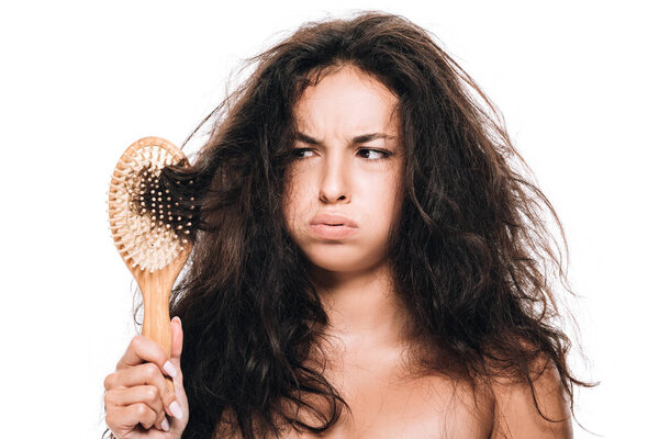 stressed brunette woman looking at wavy unruly hair in comb isolated on white