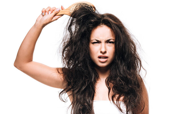 confused brunette woman styling unruly curly hair with hairbrush isolated on white