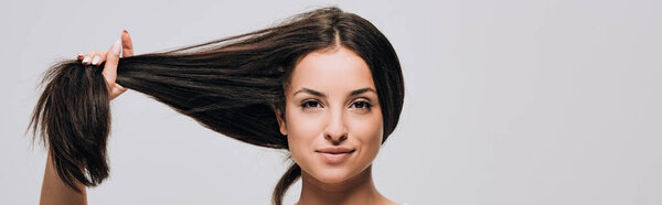 Панорамный снимок улыбающейся брюнетки красивой женщины, держащей длинные прямые здоровые и блестящие волосы, изолированные на сером
