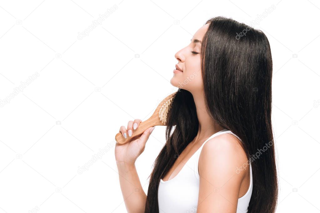side view of brunette beautiful woman with closed eyes brushing long straight healthy and shiny hair with comb isolated on white