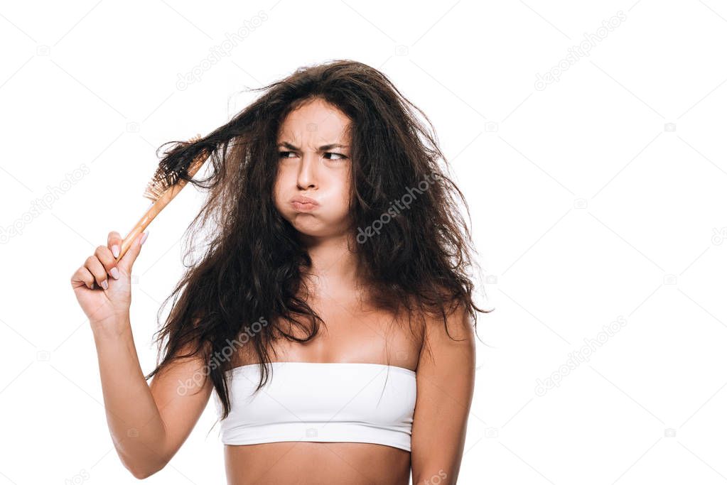 confused brunette woman looking at wavy unruly hair in comb isolated on white