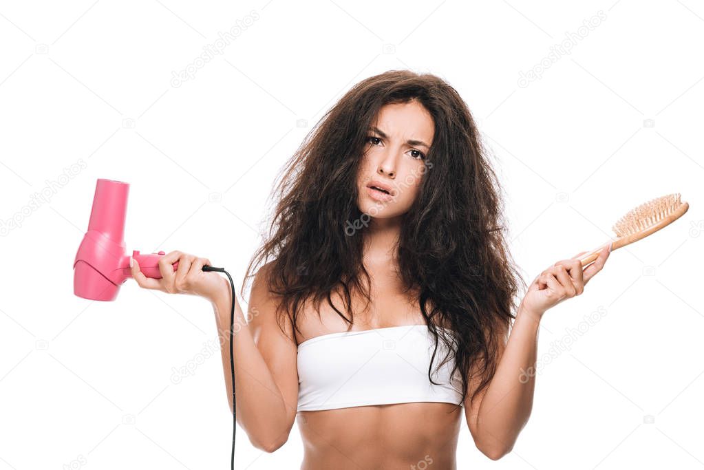 confused brunette beautiful woman with curly hair holding pink hairdryer and comb isolated on white