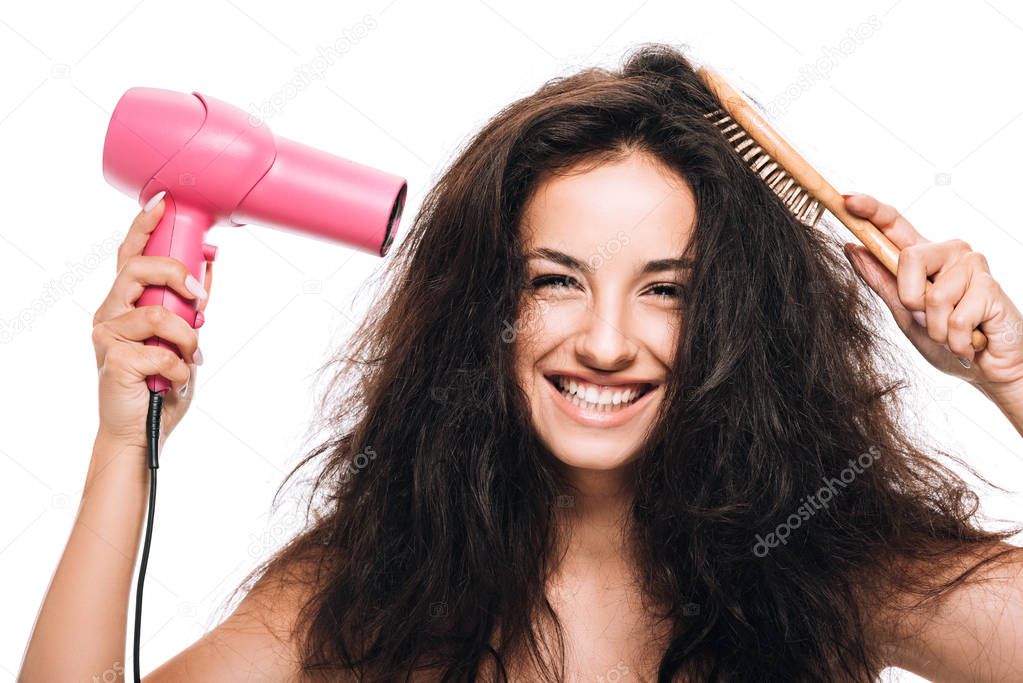 smiling brunette beautiful woman styling curly hair with pink hairdryer and comb isolated on white