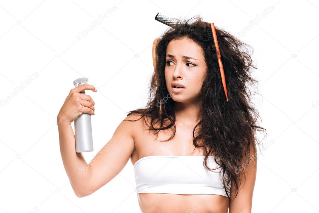 stressed brunette woman with combs in wavy unruly hair looking at spray isolated on white