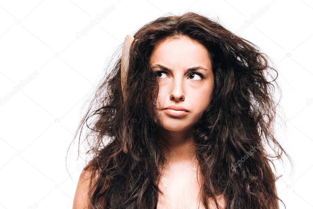 confused brunette woman with hairbrush in unruly curly hair isolated on white