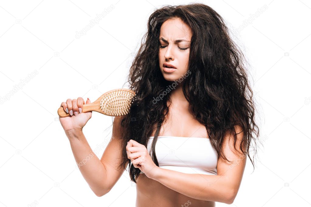 stressed brunette woman styling unruly curly hair with hairbrush isolated on white