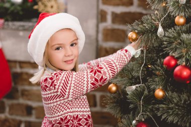 Noel Baba şapkalı ve süveterli şirin çocuk Noel ağacı süslüyor.