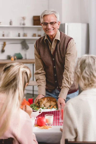 感恩节坐在桌旁的家庭成员和拿着火鸡盘子的祖父的选择焦点 — 图库照片