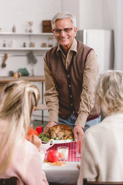 selective focus of family members sitting at table and grandfather holding plate with turkey in Thanksgiving day      