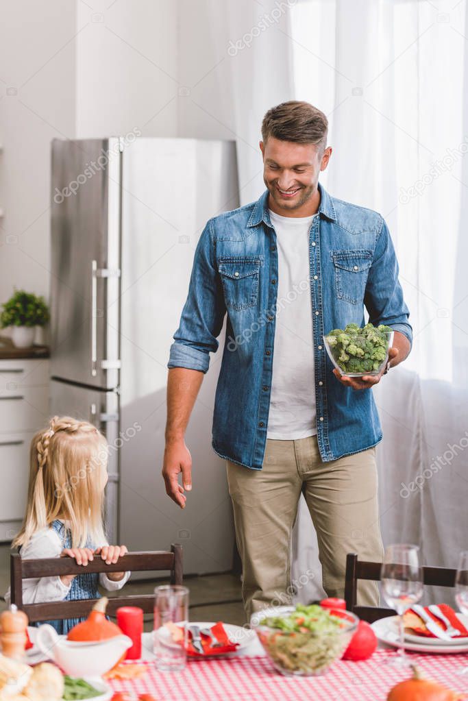 smiling father holding bowl with broccoli and looking at cute daughter in Thanksgiving day 