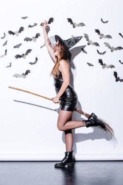 happy girl in black witch Halloween costume on broom near white wall with decorative bats clipart