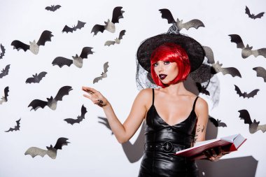 girl in black witch Halloween costume with red hair holding book near white wall with decorative bats clipart
