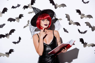 thoughtful girl in black witch Halloween costume with red hair holding book near white wall with decorative bats clipart